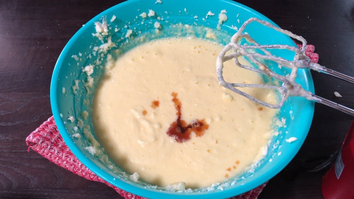 A blue bowl filled with cake batter, a splash of vanilla extract on top, and a hand mixer resting on the side.