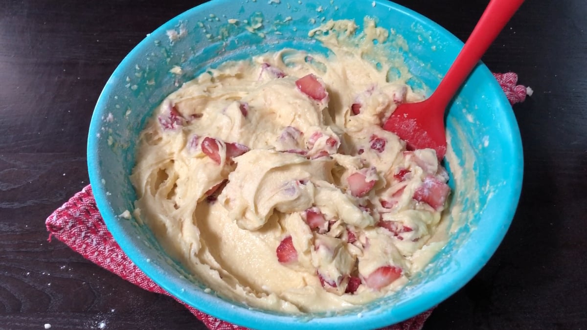 A blue bowl containing thick batter with pieces of strawberries is being mixed with a red spatula, resting on a red textured cloth on a dark surface.