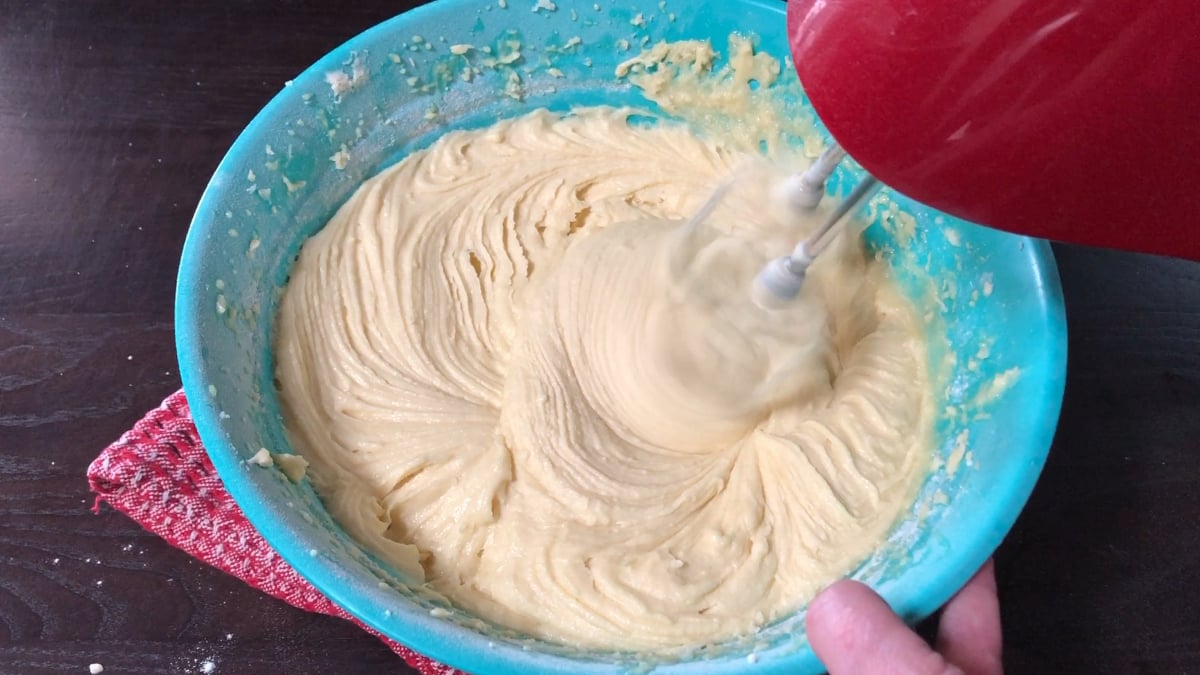 A hand holds a red electric mixer blending yellow cake batter in a blue bowl on a dark countertop.