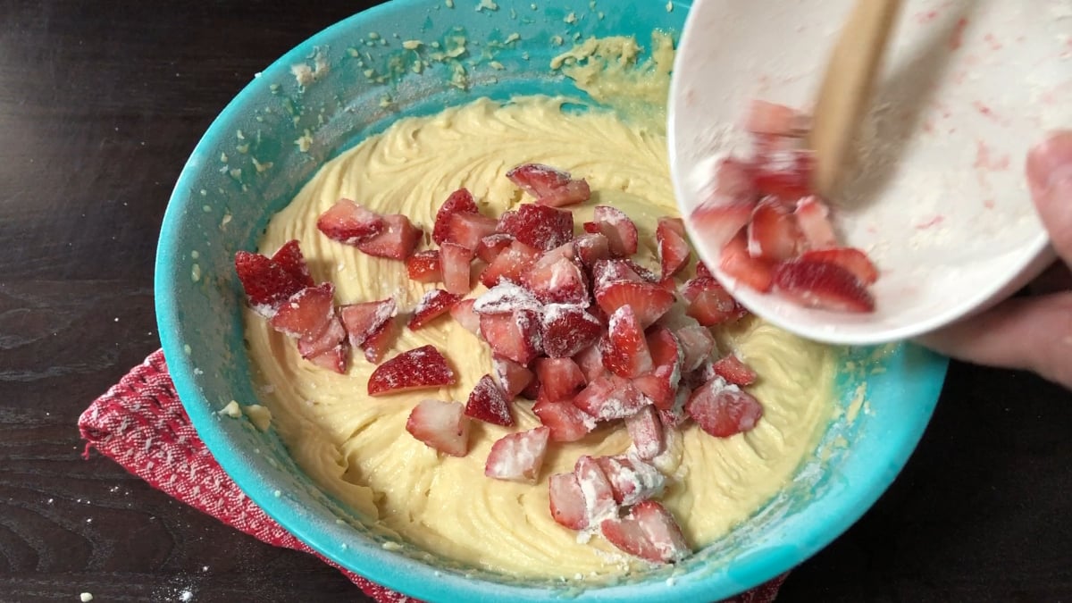 Chopped strawberries being added to a bowl of cake batter in a blue bowl, with a red textured cloth underneath on a dark surface.