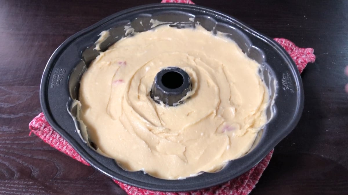 A bundt cake pan filled with unbaked cake batter rests on a red textured cloth on a dark wooden surface.