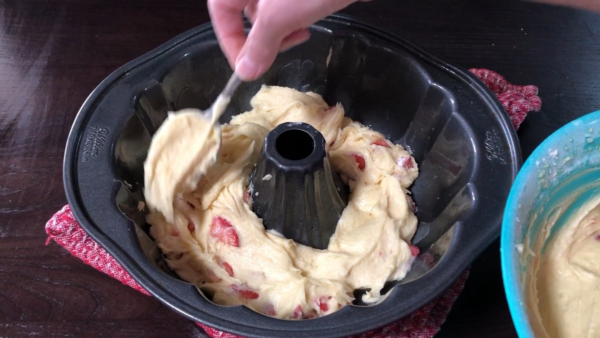 A hand spreads cake batter with pieces of fruit into a bundt pan using a spatula, with a bowl of batter and a red oven mitt nearby.