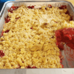 A metal baking pan with a partially served strawberry rhubarb crumble, showing a crumbly topping and a bright red fruit filling.