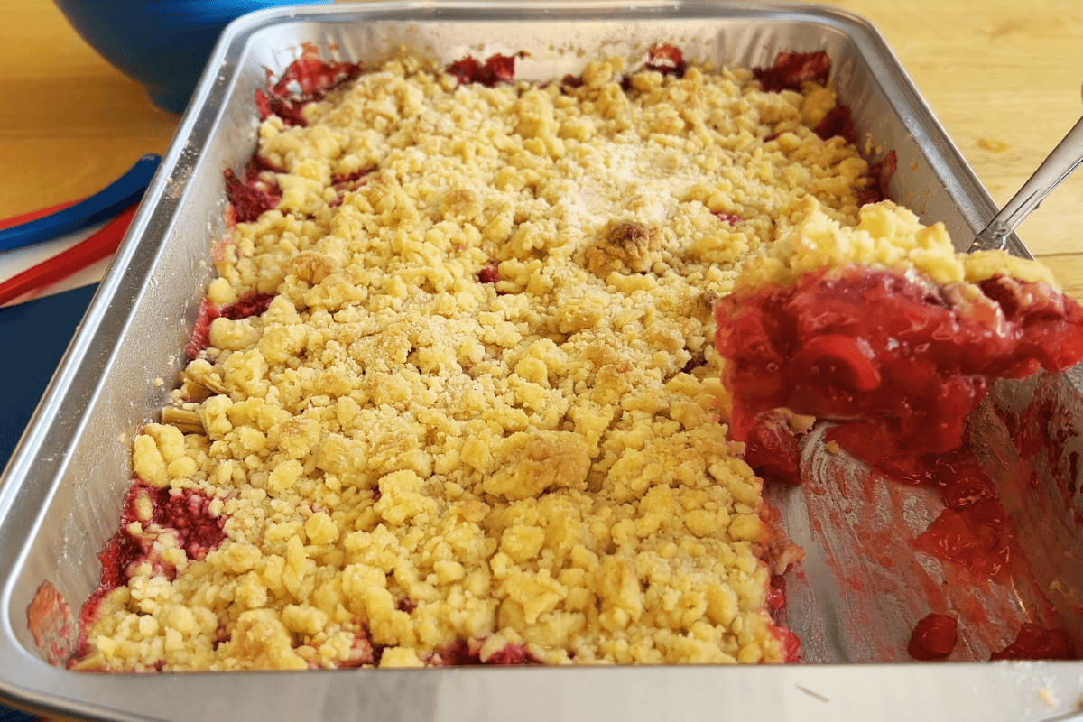 A metal baking pan with a partially served strawberry rhubarb crumble, showing a crumbly topping and a bright red fruit filling.