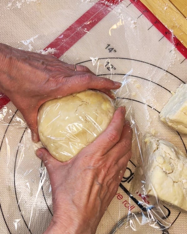 Hands wrapping a ball of dough in plastic wrap on a pastry mat, with dough scraps and a rolling pin nearby.