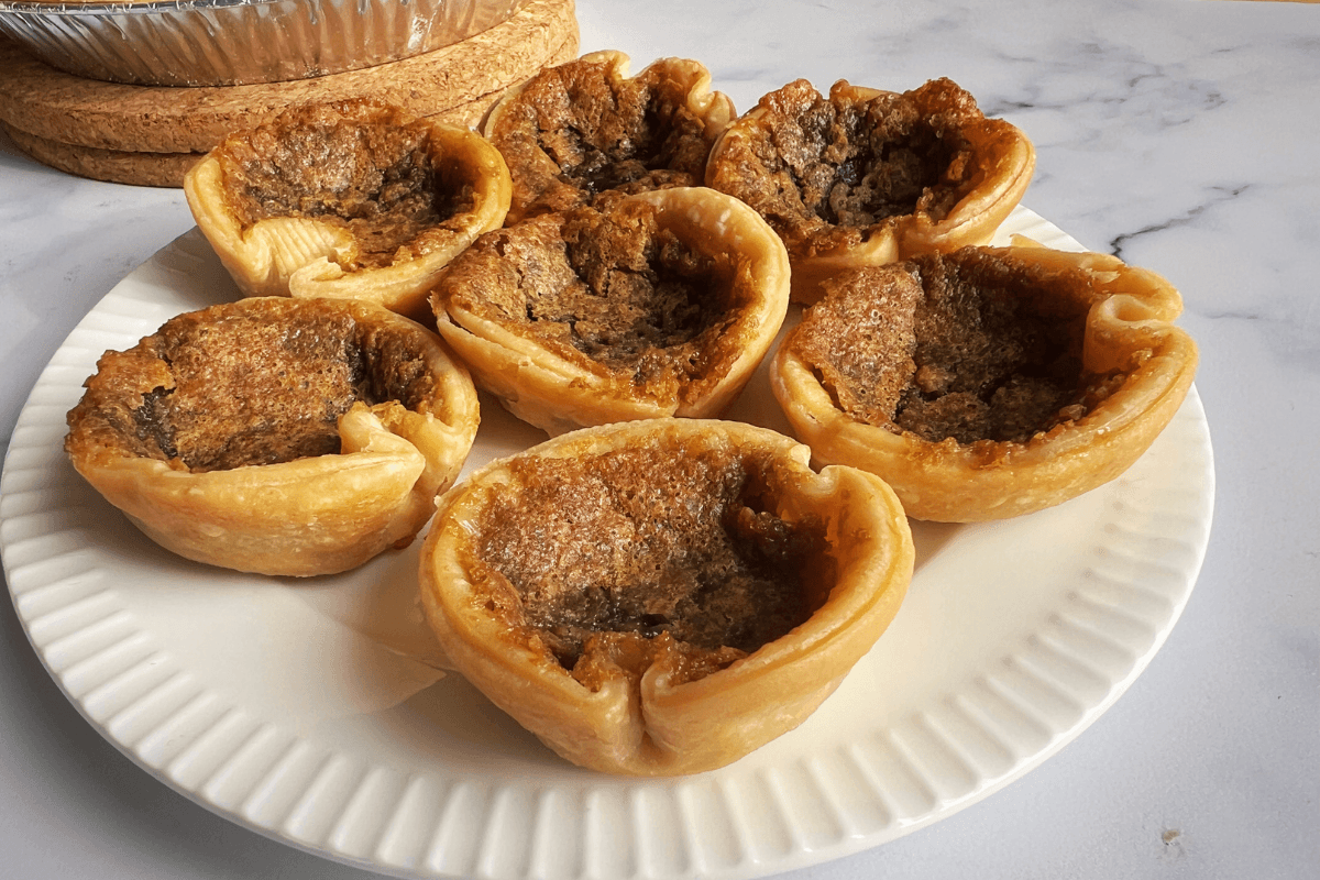 A white plate holds seven baked butter tarts with golden brown flaky crusts and caramelized filling on a marble countertop, perfect if you're looking for desserts with Pillsbury pie crust.