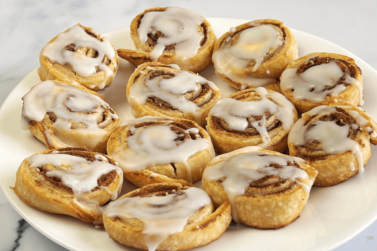 A plate of twelve cinnamon rolls topped with white icing, arranged in a single layer on a white plate—one of the easiest desserts with store bought pie crust for quick, sweet cravings.