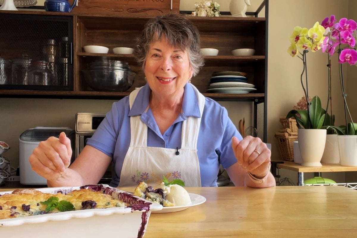An older woman in a blue shirt and white apron sits at a table with a plate of dessert, smiling. Orchids and kitchen shelves are visible in the background.