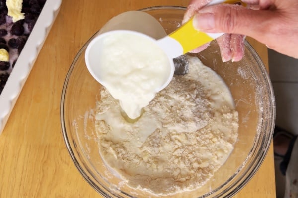 A hand pours a cup of yogurt into a bowl of flour mixture on a wooden table, with a baking dish of blueberries and butter visible nearby.
