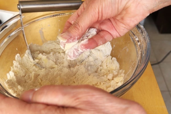 Hands mixing flour and butter in a glass bowl on a wooden surface while preparing dough.