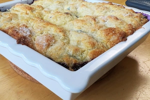 A freshly baked cobbler with a golden-brown crust sits in a white ceramic baking dish on a wooden table.