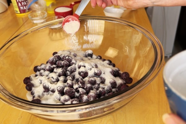 A hand sprinkles a spoonful of white powder, likely sugar or flour, over a large glass bowl filled with cherries on a wooden countertop.