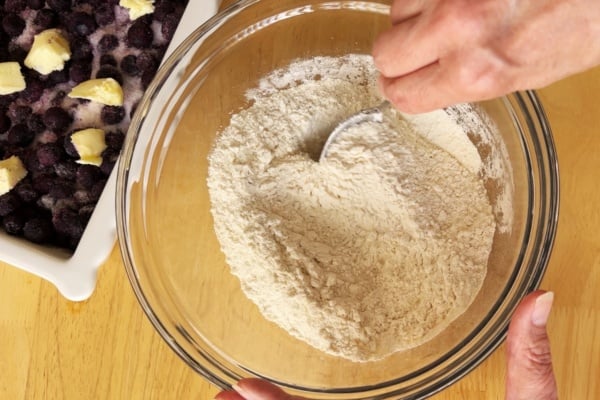 A person mixes flour with a spoon in a glass bowl next to a baking dish filled with blueberries and pieces of butter on a wooden table.