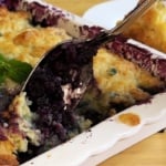 A spoon lifts a serving of blueberry cobbler from a white baking dish, showing golden-brown biscuit topping and purple filling. A sprig of mint garnishes the dessert.