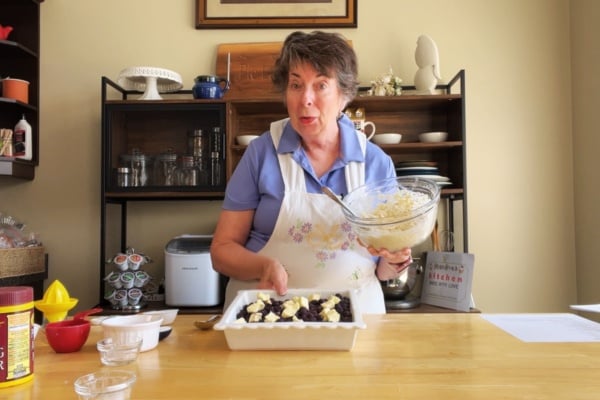 A woman in a kitchen pours a bowl of batter or topping over a dish filled with berries, preparing a dessert on a wooden table with baking tools around her.