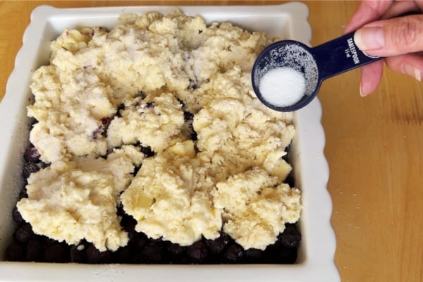 A hand sprinkles sugar from a measuring spoon over an unbaked fruit cobbler topped with biscuit-like dough in a square white baking dish.