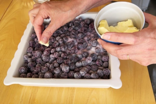 Hands place pieces of butter onto a baking dish filled with sugared frozen blueberries on a wooden table.
