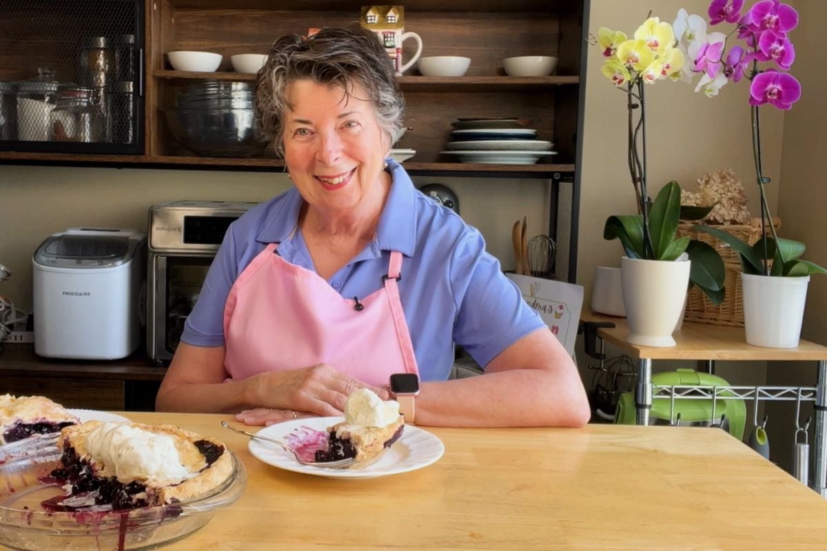 A woman in a pink apron sits at a kitchen table with a slice of pie topped with whipped cream, smiling at the camera. A whole pie and potted orchids are in the background.