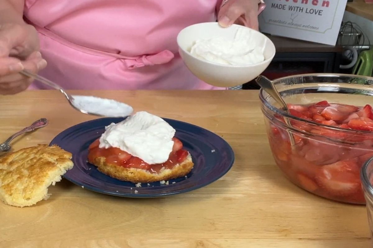 A person in a pink apron spoons whipped cream onto a strawberry shortcake on a blue plate, with sliced strawberries and more whipped cream nearby.