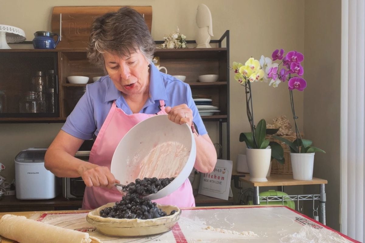 Older woman pours blueberries from a bowl into an unbaked pie crust in a home kitchen. Orchids and baking tools are visible on a table and shelf in the background.