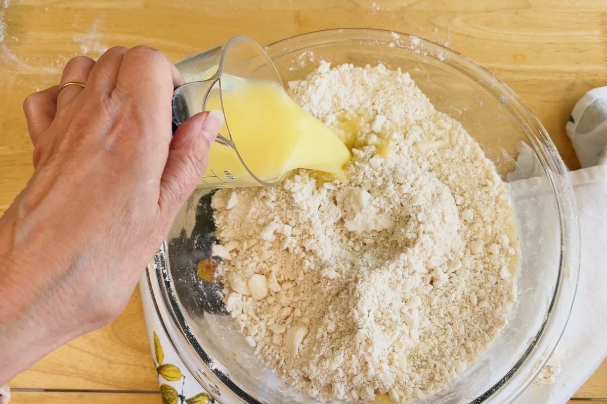 A hand pours melted butter from a measuring cup into a glass bowl filled with flour mixture on a wooden surface.