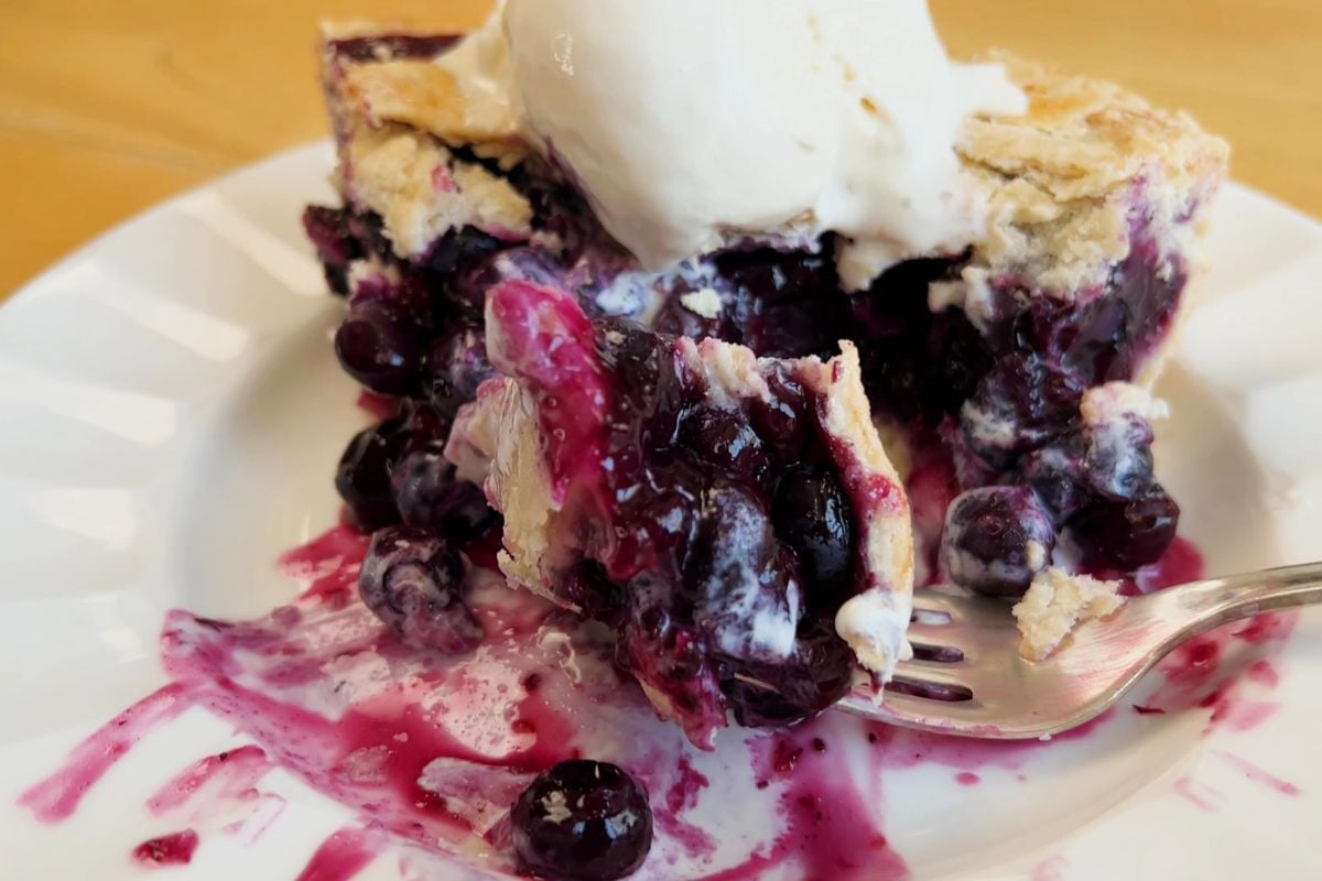 A slice of blueberry pie with a flaky crust, topped with a scoop of vanilla ice cream, sits on a white plate with a fork.