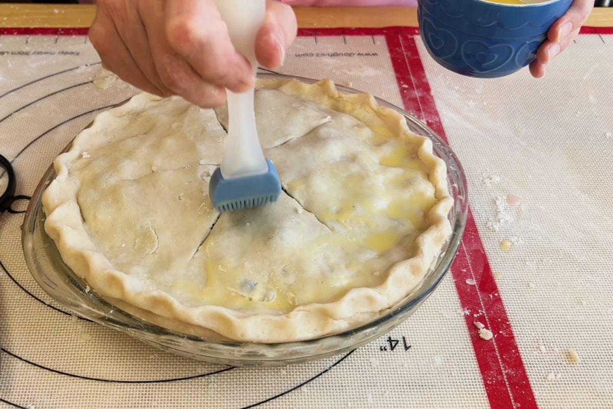 A person brushes egg wash onto an unbaked pie crust in a glass pie dish; a bowl of egg wash is held nearby on a floured pastry mat.