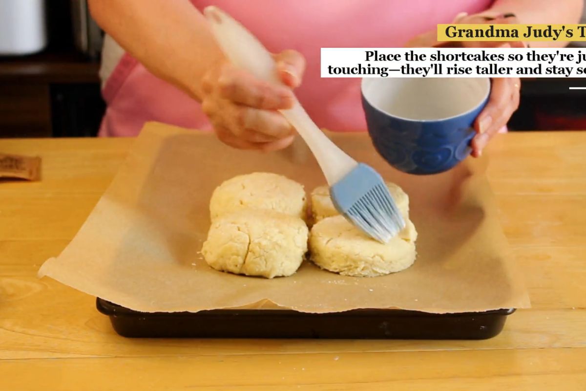 Person brushing liquid onto three unbaked shortcakes on a parchment-lined baking tray, with baking instructions partially visible in the upper right corner.