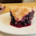 A slice of blueberry pie with a golden crust sits on a white plate, with the rest of the pie visible in the background.