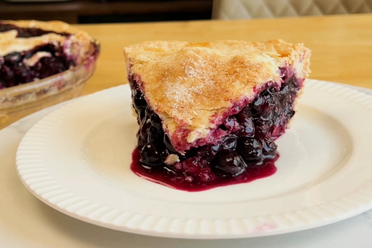 A slice of blueberry pie with a golden crust sits on a white plate, with the rest of the pie visible in the background.