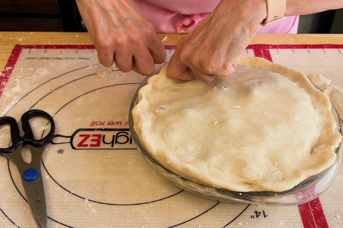 Hands crimping the edge of an unbaked pie crust in a glass pie dish on a silicone baking mat, with scissors and a marker nearby.