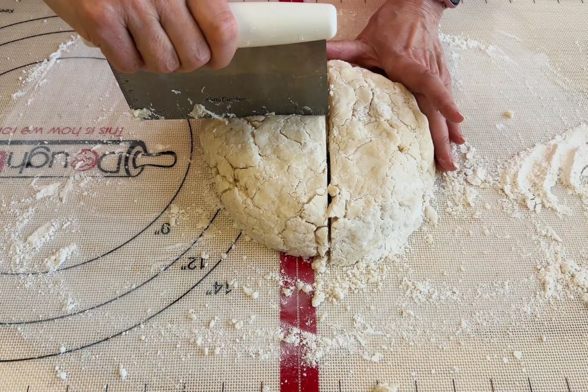 Hands use a bench scraper to cut a ball of dough in half on a floured pastry mat.