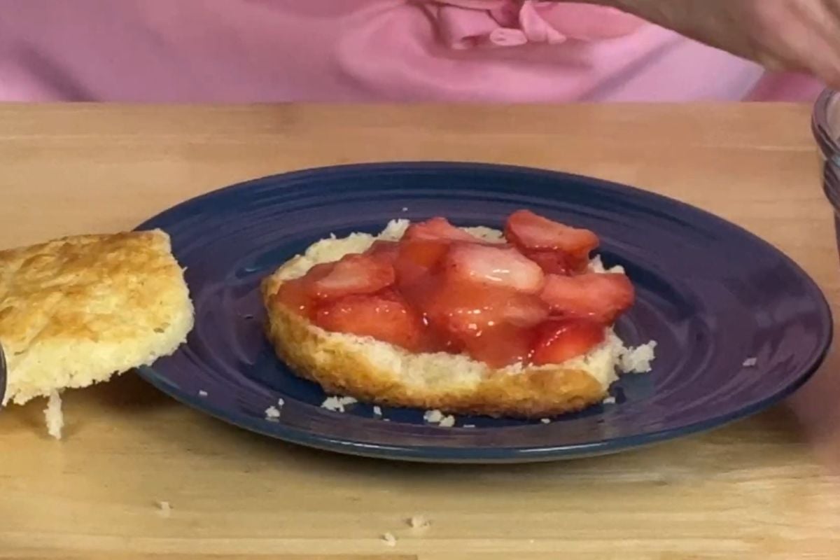 A biscuit sliced in half on a blue plate, with sliced strawberries and syrup on one half, and the other half set to the side.