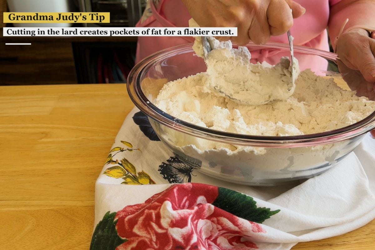 A person uses a spoon to cut lard into flour in a glass bowl on a wooden table covered with a floral cloth. Text overlay shares a baking tip about making a flakier crust.