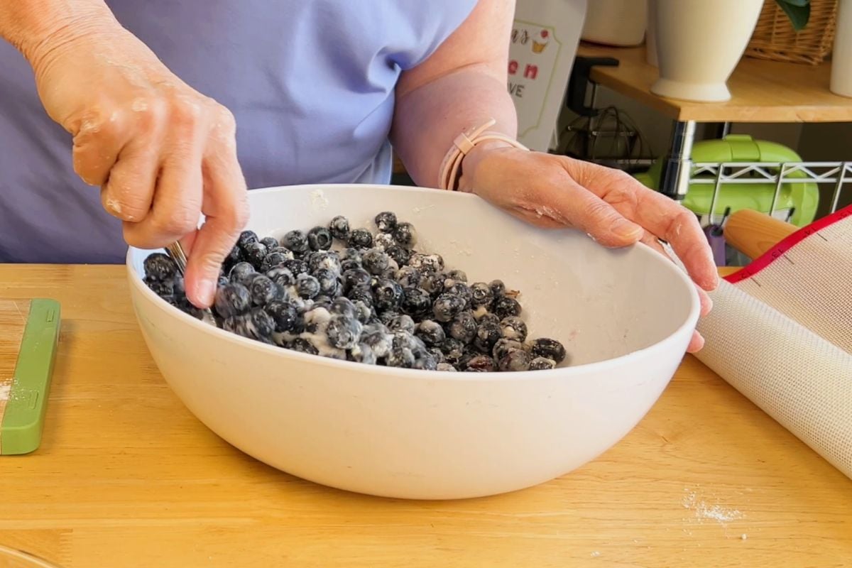 A person mixes blueberries and flour in a large white bowl on a wooden table, preparing ingredients for baking.