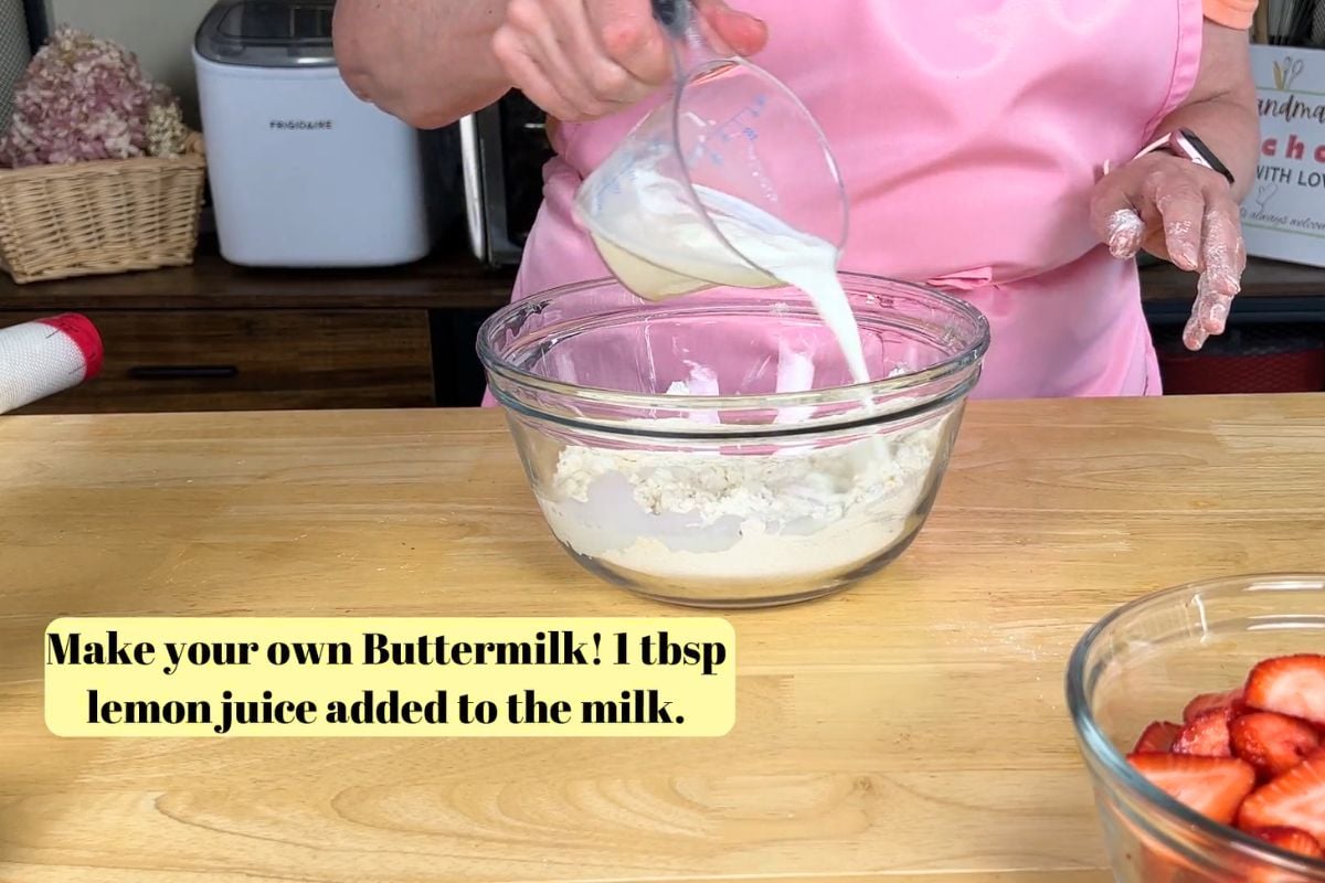 A person in a pink apron pours milk into a glass bowl with flour on a wooden counter. A bowl of sliced strawberries sits nearby. Text on the image gives a buttermilk recipe.