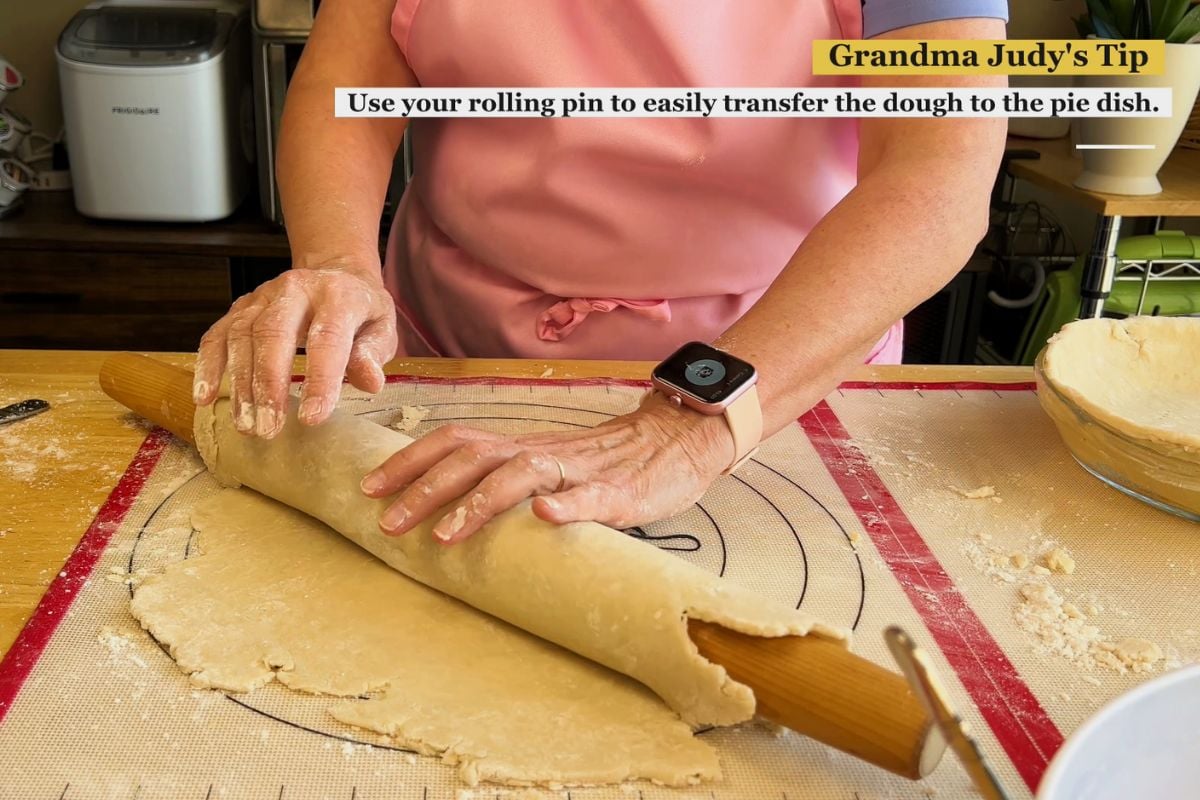 Person in a pink apron rolls pie dough onto a wooden rolling pin on a floured surface, preparing to transfer it. Text overlay gives a tip about using a rolling pin for dough transfer.
