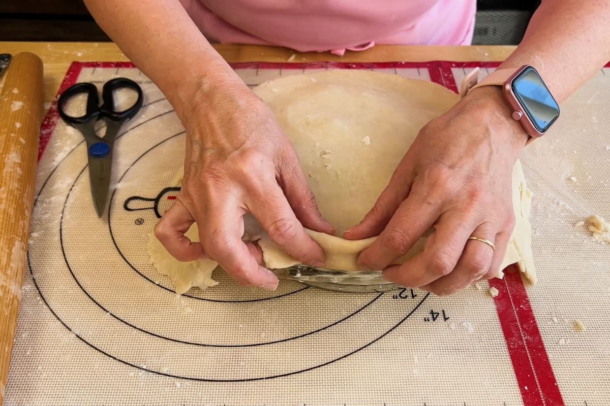 Person folding pie dough over the edge of a pie dish on a pastry mat, with a watch on their wrist and scissors nearby.