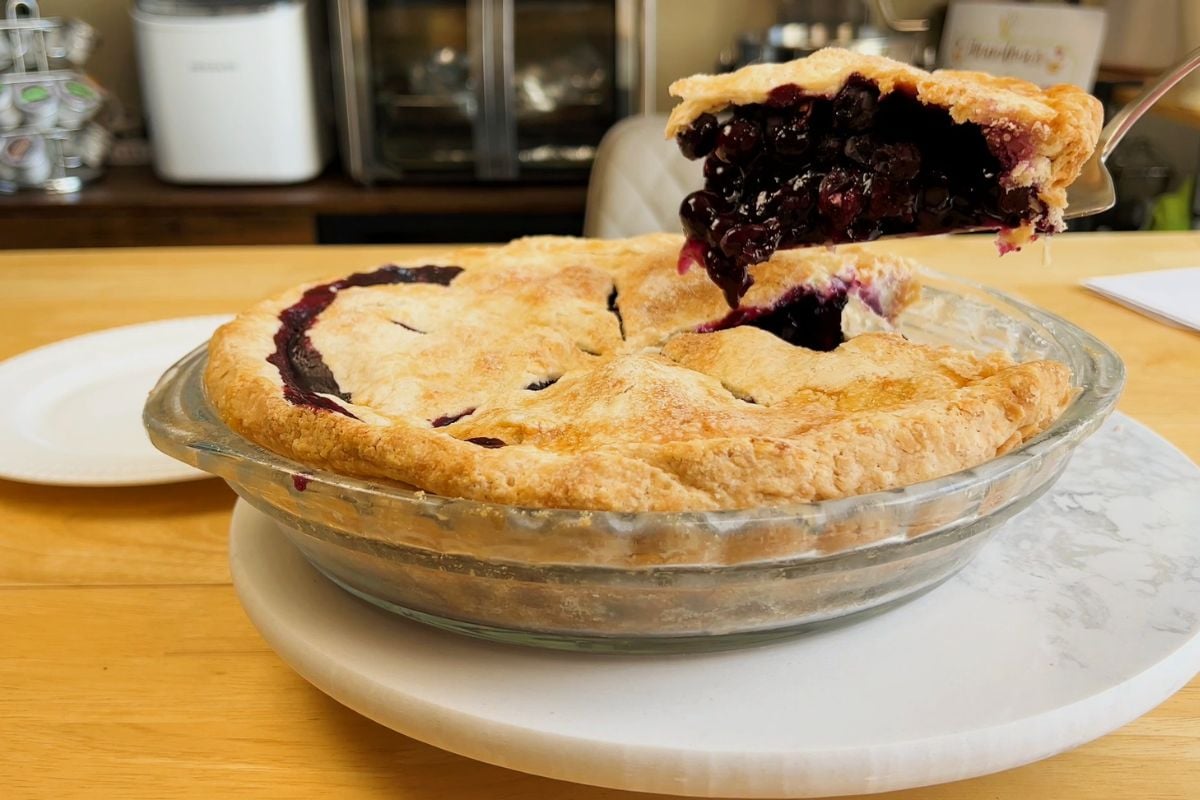 A slice of blueberry pie is being lifted from a glass pie dish, showing a golden crust and dark filling, on a wooden countertop.
