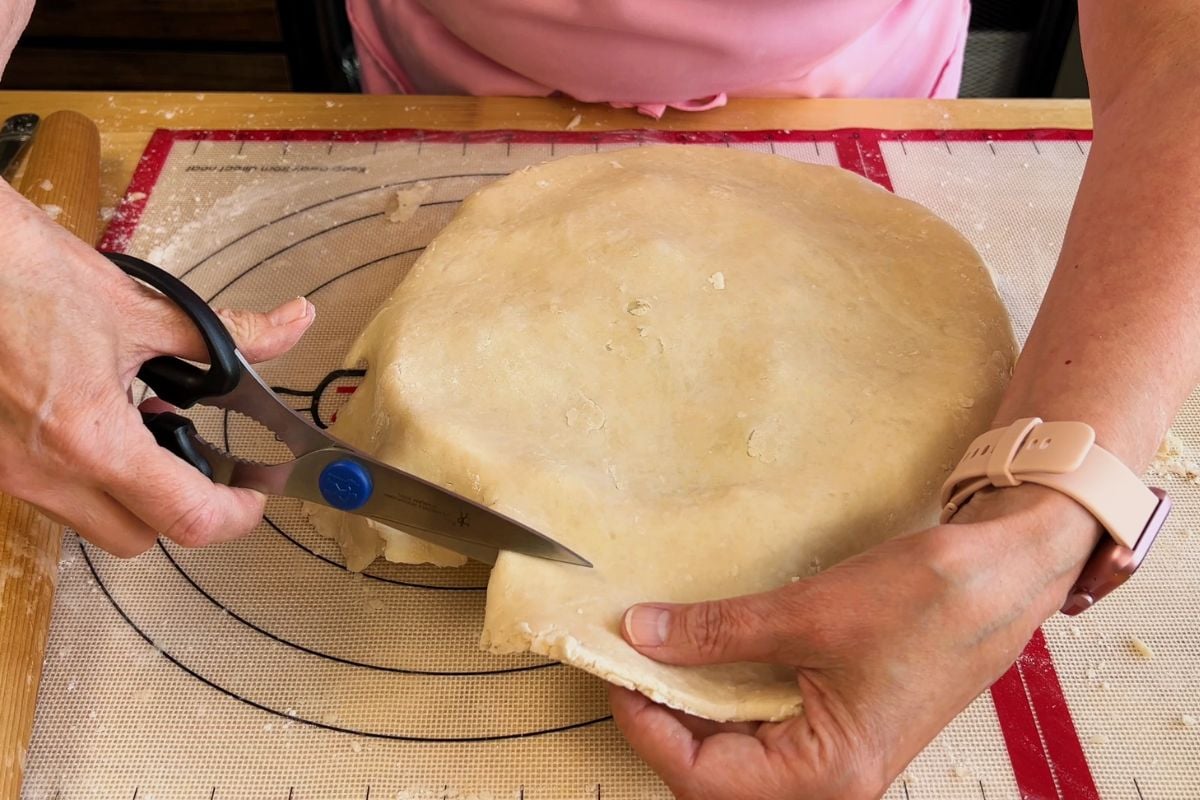 A person uses kitchen scissors to trim excess dough from the edge of a rolled pie crust on a silicone baking mat.