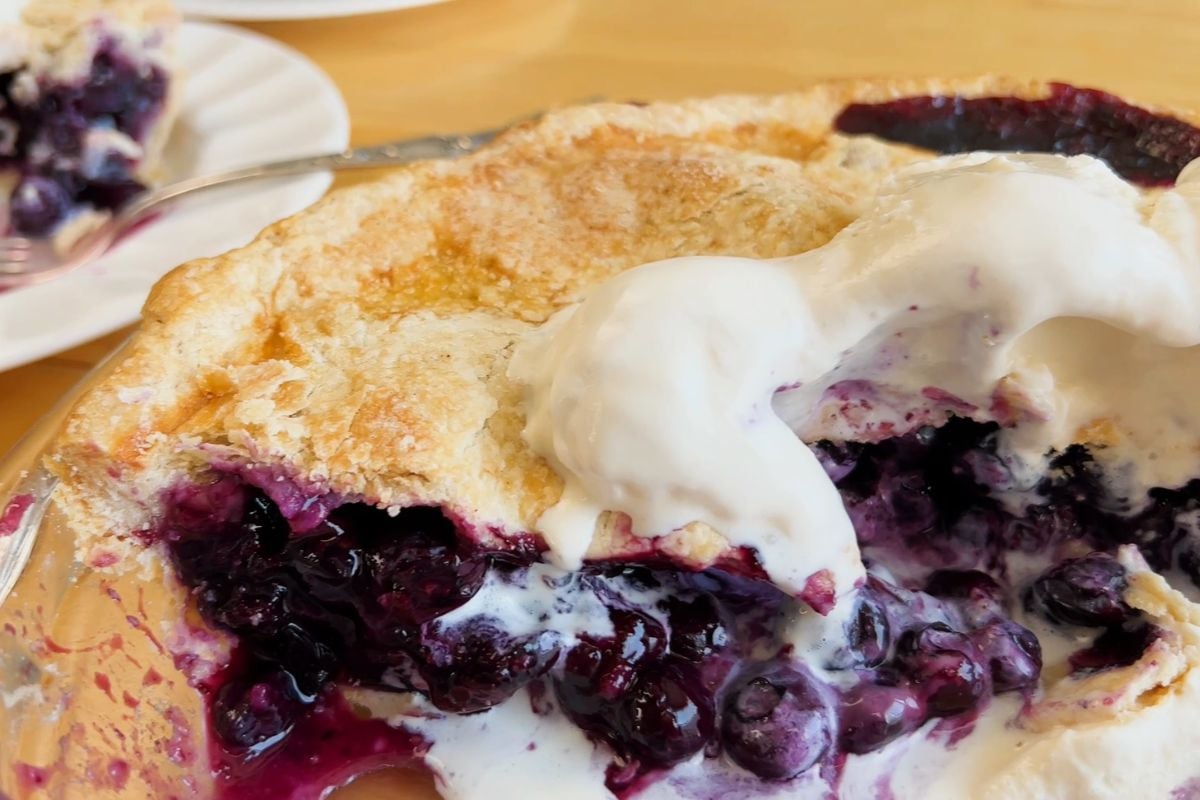 A partially eaten blueberry pie topped with a scoop of melting vanilla ice cream, with a plate holding a slice in the background.